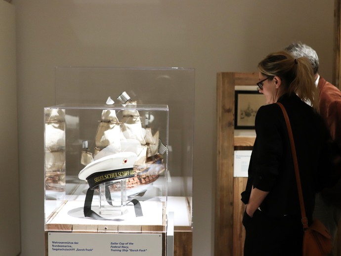 Eine Besucherin betrachtet eine Vitrine, in der sich eine Matrosenmütze befindet. (vergrößerte Bildansicht wird geöffnet)