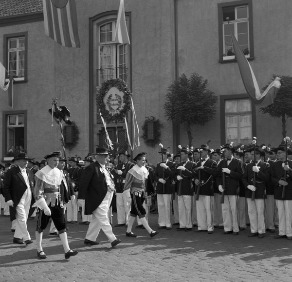 Auf der Schwarzweißfotografie sieht man einen Festumzug durch die Innenstadt. Die Anwesenden tragen traditionelle Kleidung und Uniformen.